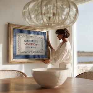 May include: A framed artwork with a gold-colored frame. The artwork features the text "GOD BLESS AMERICA" with stars and the years "1776 - 2026" on a blue and white gingham background. Floral and leaf patterns surround the text.