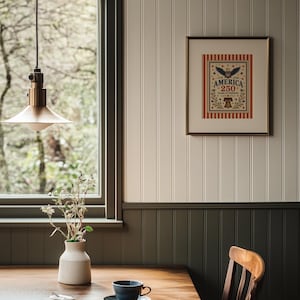Puede incluir: Interior de comedor con mesa de madera, taza y platillo azules y obra de arte enmarcada con el texto "AMERICA 250 YEARS OF FREEDOM". Un jarr&oacute;n blanco con flores secas est&aacute; sobre la mesa. Una l&aacute;mpara colgante de lat&oacute;n cuelga encima.