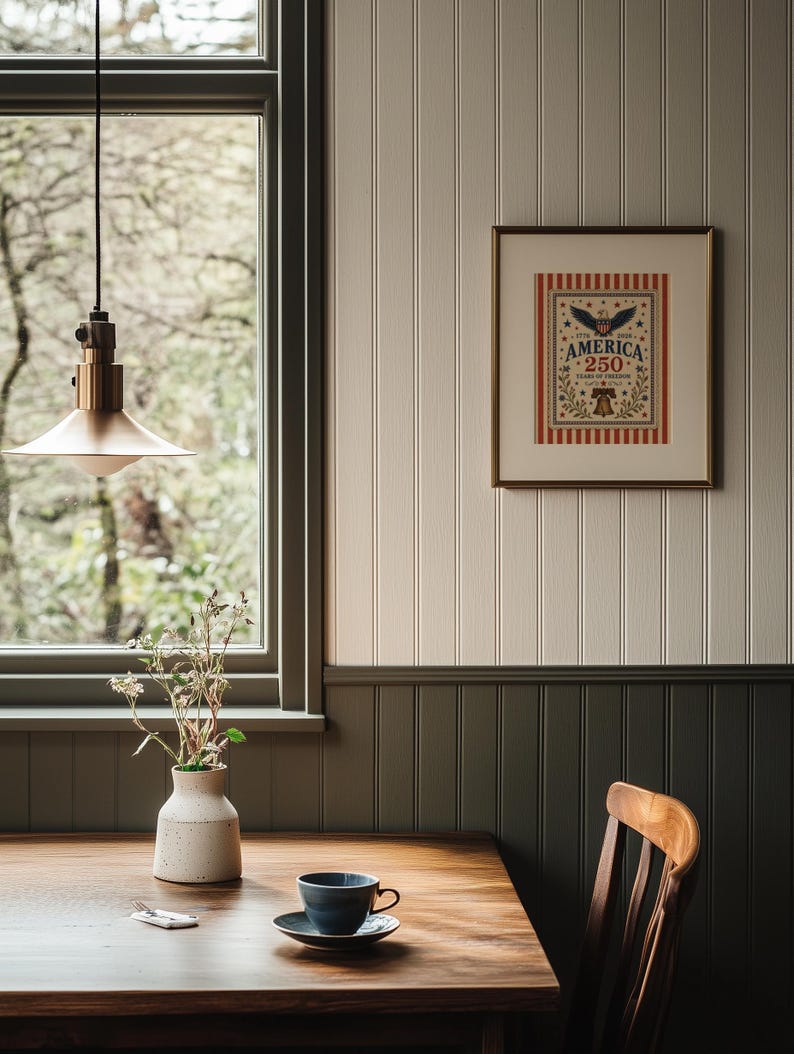 Puede incluir: Interior de comedor con mesa de madera, taza y platillo azules y obra de arte enmarcada con el texto "AMERICA 250 YEARS OF FREEDOM". Un jarr&oacute;n blanco con flores secas est&aacute; sobre la mesa. Una l&aacute;mpara colgante de lat&oacute;n cuelga encima.