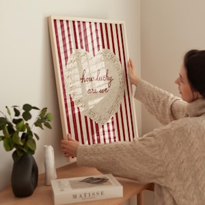 May include: A framed artwork with a red and white striped background. A heart-shaped doily with the words "how lucky are we" is centered on the artwork. The frame is white. The artwork is being held by a person.