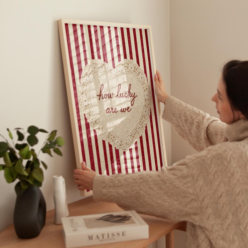 May include: A framed artwork with a red and white striped background. A heart-shaped doily with the words "how lucky are we" is centered on the artwork. The frame is white. The artwork is being held by a person.