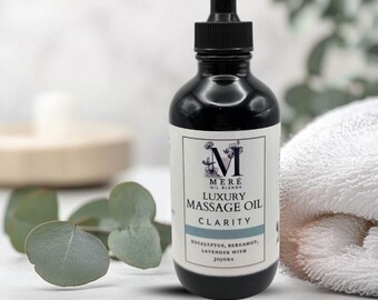 May include: A black glass bottle of "Luxury Massage Oil Clarity" with a dropper. The label includes the brand name "Mere Oil Blends" and lists ingredients like eucalyptus, bergamot, and lavender. A white towel and eucalyptus leaves are in the background.