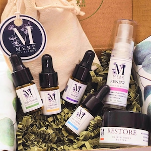 May include: A gift box filled with a variety of essential oil blends and body care products. The box includes a small bag, four glass bottles with droppers, a bottle of hair and body gel, and a jar of hair and body butter. The products are labeled with the brand name "Mere Oil Blends" and feature different scents and uses.