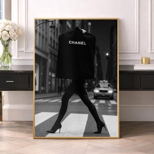 Könnte beinhalten: Ein schwarz-weiß gerahmter Wandkunstdruck, der eine Person zeigt, die über einen Zebrastreifen geht und eine schwarze Kleiderhülle mit dem weißen Schriftzug CHANEL trägt. Die Person trägt eine schwarze Strumpfhose und schwarze High Heels.