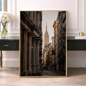 May include: A framed vertical wall art print featuring a street-level perspective of a New York City avenue. The composition highlights tall buildings with fire escapes lining the street, leading the eye toward the Empire State Building in the distance.