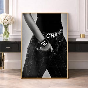 Könnte beinhalten: Ein schwarz-weißes Wandbild, das einen Oberkörper in einem schwarzen Oberteil und einer dunklen Jeans mit einem Gürtel mit der Aufschrift CHANEL zeigt. Ein Armband mit dem ineinandergreifenden CC-Logo ist am Arm in der Tasche zu sehen.