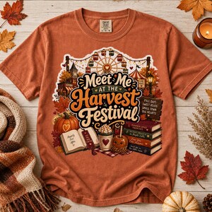 Könnte beinhalten: Ein rostfarbenes T-Shirt mit einem Grafikdruck eines Erntefestes, einem Riesenrad, einem Kürbis, einem offenen Buch, einem Bücherstapel und einem kandierten Apfel. Der Text lautet Meet Me at the Harvest Festival und Fall Days.
