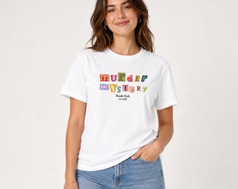 May include: A white short-sleeved t-shirt featuring the text "Murder Mystery" in colourful, ransom-note style letters across the chest, with the words "Book Club" and "est 2016" printed in black underneath the main graphic.