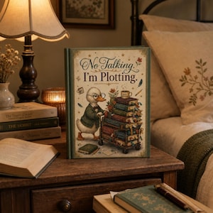 May include: A hardcover book with a cream-colored cover featuring a duck wearing glasses and a green jacket, pushing a cart filled with stacked books. The text on the cover reads, No Talking. I'm Plotting. The book has a dark green border and gold accents.