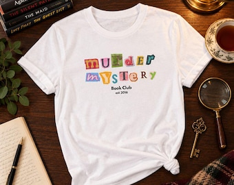 Op de afbeelding: Een wit t-shirt met korte mouwen voorzien van de tekst "murder mystery" in een kleurrijk lettertype in losse-letterstijl, met de woorden "Book Club" en "est 2016" in het zwart gedrukt onder de hoofdafbeelding op het midden van de borst.