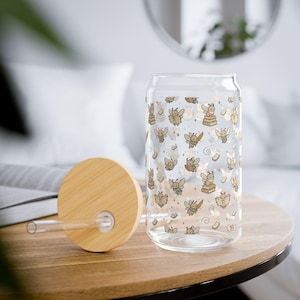 Könnte beinhalten: Ein durchsichtiges Glas mit einem Bambusdeckel und einem Glasstrohhalm. Das Glas ist mit einem sich wiederholenden Muster aus kleinen, beigefarbenen Feenfiguren, Büchern und winzigen Sternen bedruckt. Die Feen sind in verschiedenen Posen dargestellt.