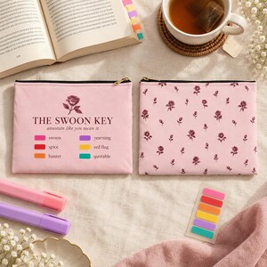 May include: Two rectangular light pink zippered pouches. One pouch features a dark rose illustration, the text THE SWOON KEY, and a color-coded legend with terms like swoon, spice, banter, yearning, red flag, and quotable. The other has a rose pattern.