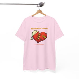 May include: A light pink short-sleeve t-shirt featuring a graphic of a red heart with a bandage, stitches, and a small adhesive bandage. The text "Enemies to Lovers" appears above the heart, and "I Tolerated You... Still I Didn't" is below.