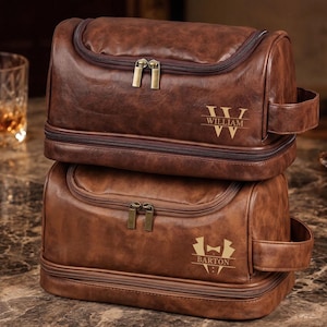 Op de afbeelding: Twee gestapelde bruine lederen toilettassen met donkerbruine ritsen en handvatten aan de zijkant. De bovenste tas toont de naam WILLIAM met een W-monogram, en de onderste tas toont de naam BARTON met een smoking-afbeelding, beide in goudkleurige letters.