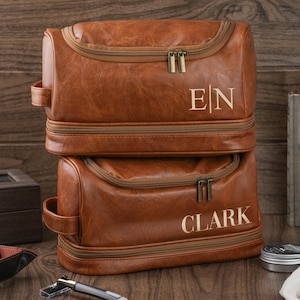 May include: Two stacked brown leather toiletry bags with gold zippers and handles. The top bag has the initials "EN" in gold, and the bottom bag has the name "CLARK" in gold. The bags are on a wooden surface.