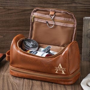 May include: A brown leather travel toiletry bag with a zippered top and a separate compartment. The bag is monogrammed with the name "Ahern" in gold lettering.