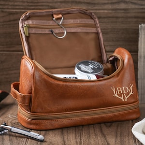 May include: A brown leather travel toiletry bag with a zippered top and a separate compartment. The bag has a monogrammed design with the initials "BD" and a pair of antlers.
