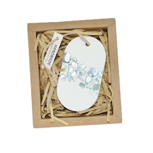 May include: A white ceramic oval ornament with a blue and silver mosaic design. The ornament is in a brown cardboard box with shredded paper. The ornament has a tag that says "Handmade with love for you".