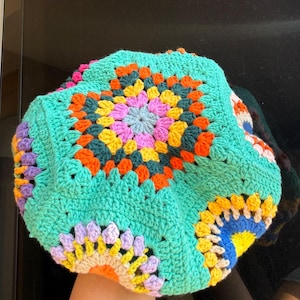 Op de afbeelding: Een gehaakte muts met een levendig granny square patroon en een centraal motief in tinten turquoise, oranje, geel, roze en paars. Het ontwerp bestaat uit meerdere met elkaar verbonden vierkanten met een opvallende turquoise rand.