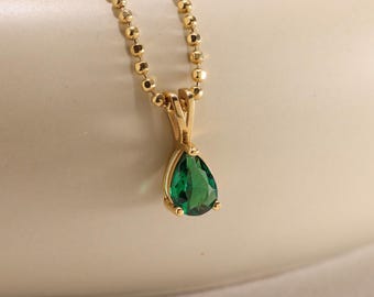 Puede incluir: Un collar dorado con un colgante en forma de lágrima de color verde esmeralda. El colgante cuelga de una delicada cadena con pequeñas cuentas redondas. El collar se exhibe sobre una superficie curva de color crema.