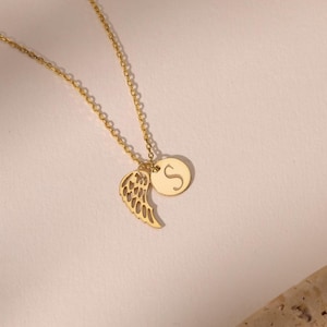 May include: A gold-colored necklace with two pendants. One pendant is a wing shape, and the other is a small, round disc with the letter "S" on it. The necklace is on a light-colored background.