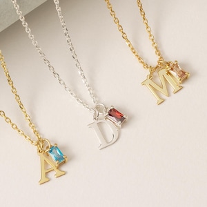May include: A selection of initial pendant necklaces. The necklaces are gold and silver, with the letters "A", "D", and "M" displayed. Each letter pendant is paired with a gemstone, including blue and red. The chains are delicate.