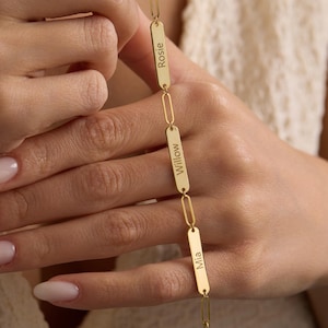 May include: A gold-colored bracelet with a chain link design. The bracelet features three rectangular nameplates engraved with the names "Rosie", "Willow", and "Mia". The bracelet is held up by a person.