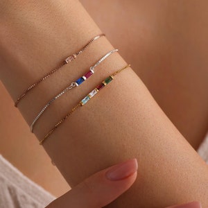 May include: Four delicate bracelets with colorful gemstones on a woman's wrist. The bracelets are made of gold and silver chains with small, square-shaped gemstones in shades of blue, green, red, and pink.