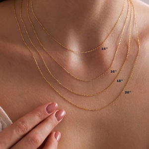 May include: Four delicate gold necklaces of varying lengths, displayed on a person's chest. The necklaces are labeled with their lengths: 14", 16", 18", and 20". The chains are fine and elegant, showcasing a minimalist jewelry style.