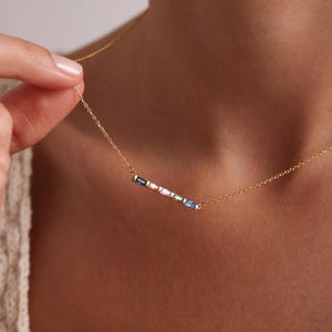 May include: A gold chain necklace with a delicate bar pendant featuring a row of colorful gemstones. The gemstones are arranged in a rainbow pattern, with shades of blue, pink, and green.