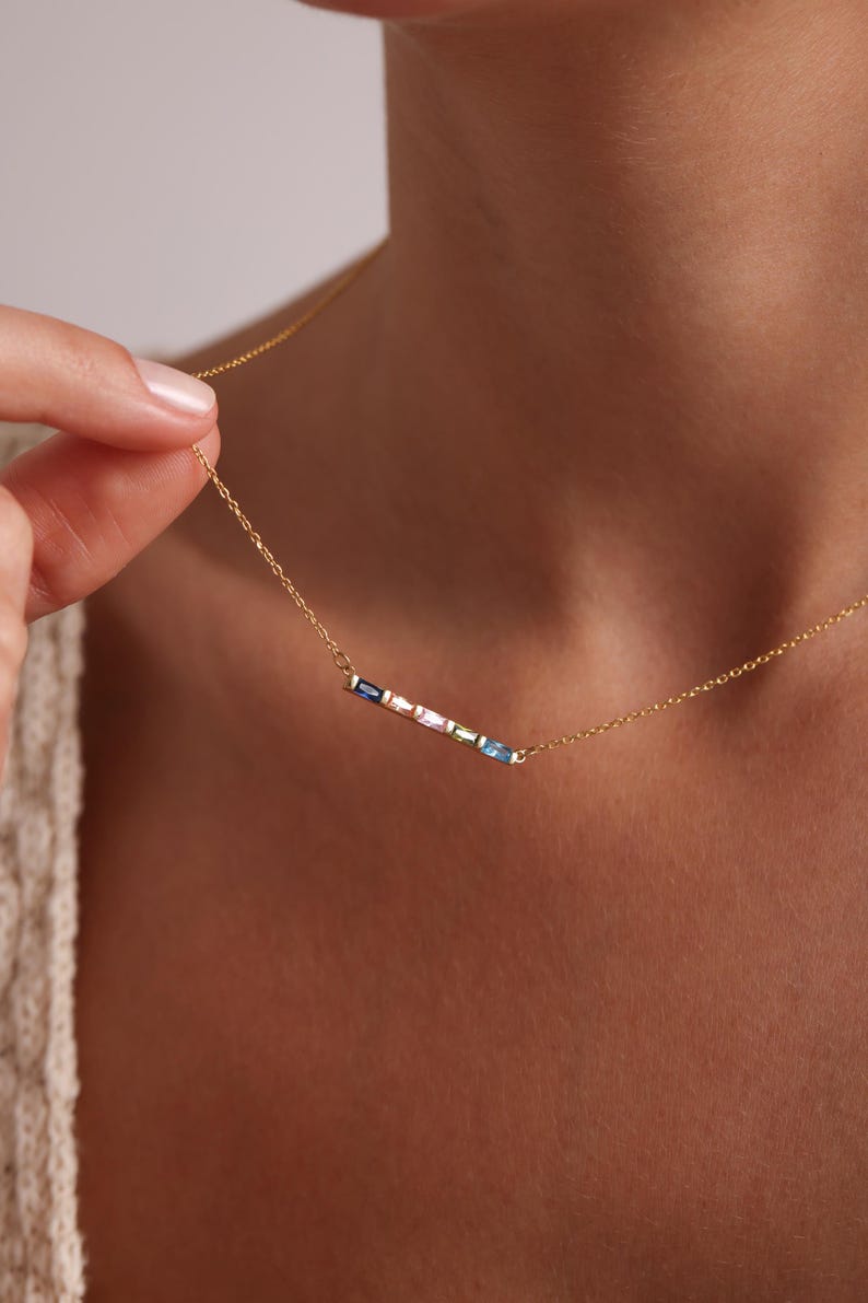 May include: A gold chain necklace with a delicate bar pendant featuring a row of colorful gemstones. The gemstones are arranged in a rainbow pattern, with shades of blue, pink, and green.
