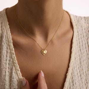 May include: A delicate gold necklace featuring a heart-shaped pendant with a wing design and a small circular charm with the letter "S". The necklace is worn around the neck, showcasing its intricate details and elegant design.