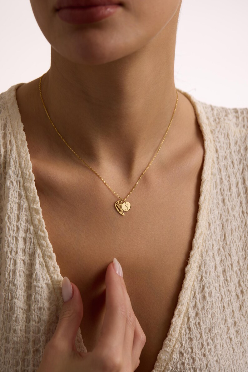 May include: A delicate gold necklace featuring a heart-shaped pendant with a wing design and a small circular charm with the letter "S". The necklace is worn around the neck, showcasing its intricate details and elegant design.