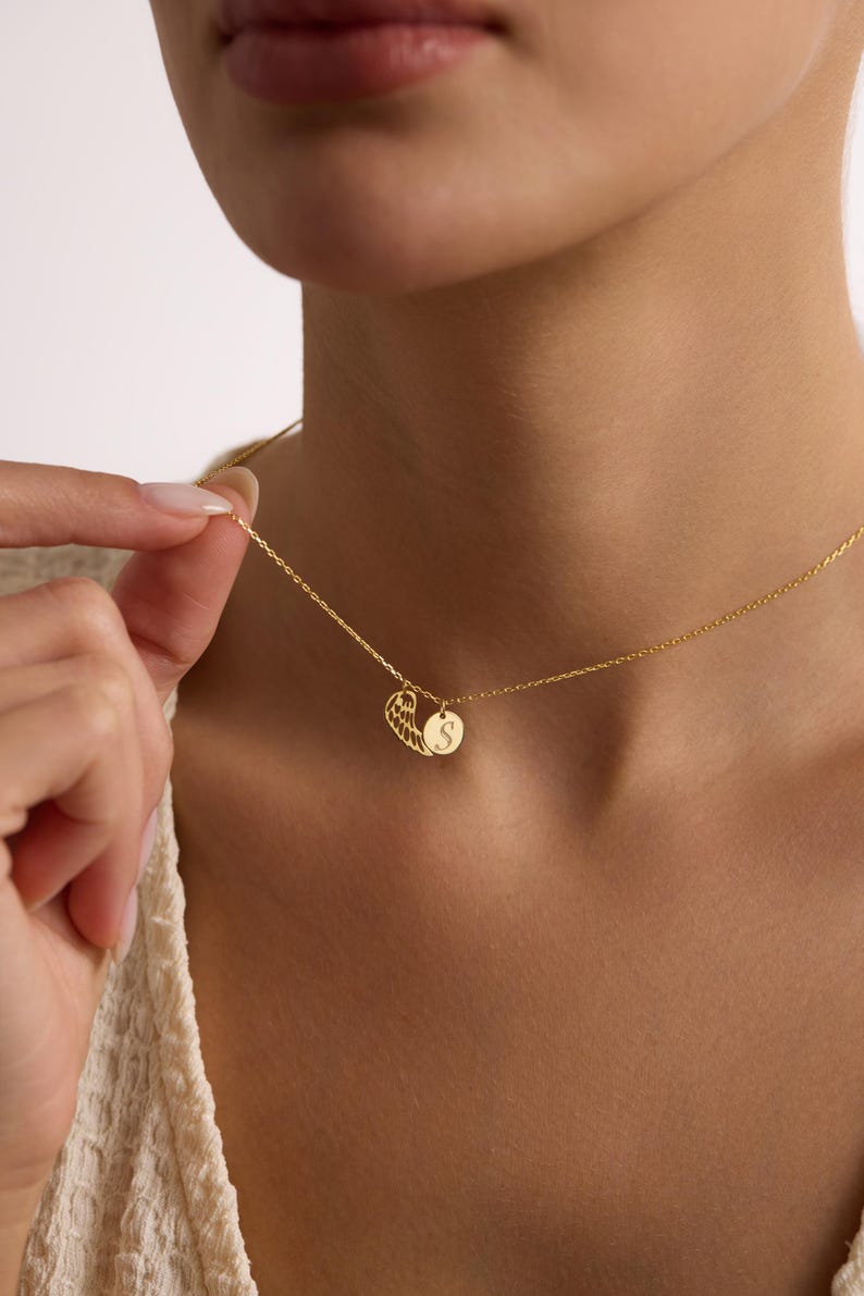 May include: Gold-colored necklace with two pendants. One pendant is a wing design, and the other is a small, round charm with the letter "S" engraved on it. The necklace is delicate and appears to be made of a thin chain.
