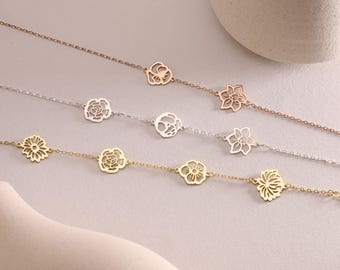 May include: Four delicate gold chains with different flower-shaped charms. The chains are arranged in a row on a light beige surface.