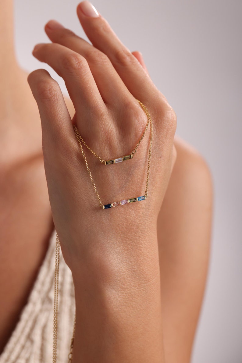 May include: Two gold necklaces with delicate chains and colorful gemstone bar pendants. The top necklace features a single bar with clear gemstones, while the bottom necklace has a bar with multiple colorful gemstones.