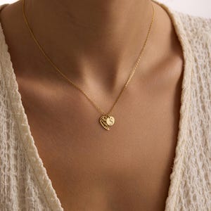 May include: A gold-colored necklace featuring a delicate wing charm and a round pendant with the letter "S". The necklace is worn against a cream-colored top, creating a subtle contrast. The chain is fine and elegant.