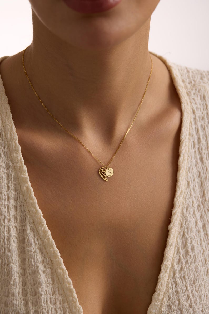 May include: A gold-colored necklace featuring a delicate wing charm and a round pendant with the letter "S". The necklace is worn against a cream-colored top, creating a subtle contrast. The chain is fine and elegant.