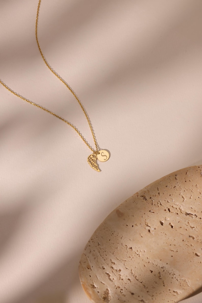 May include: A gold-colored necklace with two pendants. One pendant is a wing shape, and the other is a small, round disc with the letter "S" on it. The necklace is on a light-colored background.