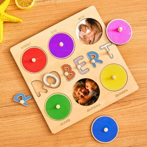 Peut inclure: Puzzle en bois avec le nom "ROBERT" et des découpes circulaires pour des photos. Le puzzle comporte des cercles rouges, violets, verts, jaunes, roses et bleus. Le mot "ROBERT" est épelé avec des lettres individuelles. Le puzzle comprend les mots "MOM", "DAD", "GRANDPA", "GRANDMA" et "OLIVER".