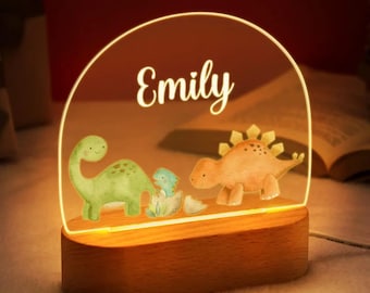Puede incluir: Una luz de noche personalizada con una base de madera y un panel acrílico transparente arqueado. El panel muestra el nombre "Emily" en escritura elegante sobre ilustraciones de acuarela de dinosaurios en verde, naranja y azul.