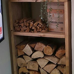 Könnte beinhalten: Ein hohes, rechteckiges Holzregal mit zwei Ebenen. Das untere Fach ist mit gestapeltem Brennholz gefüllt. Das obere Fach enthält Anzündholz, eine Schachtel mit der Aufschrift 'Hotblaze FIRELIGHTERS', einen Bilderrahmen und eine hängende Grünpflanze.