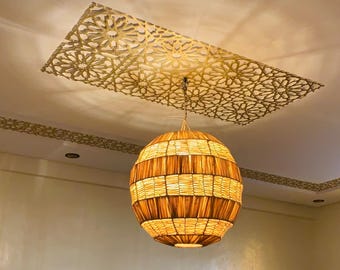 May include: A spherical pendant light made of woven material hangs from the ceiling, emitting a warm, amber light. Above, a rectangular panel with intricate cut-out patterns adds a decorative element. The ceiling and walls are a soft, neutral colour.