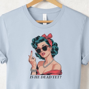 May include: Light blue t-shirt with a graphic of a woman wearing sunglasses and a red bow in her hair. The woman is giving the middle finger and the text "Is he dead yet?" is printed below the graphic.