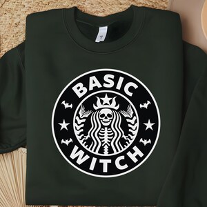 May include: A dark forest green crewneck sweatshirt featuring a white circular graphic on the chest. The design depicts a skeletal figure with long hair and a crown, surrounded by small bats and stars, with the text BASIC WITCH printed in white.