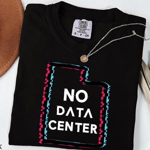 May include: Black t-shirt with the text "NO DATA CENTER" in white, framed by a glitch-style design in pink and blue. A silver necklace with a small pendant rests on the shirt. The shirt is a Comfort Colors brand.