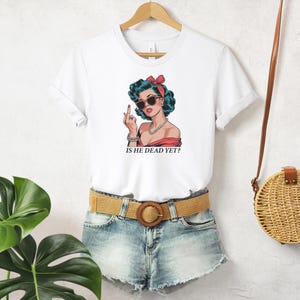 May include: White t-shirt with a cartoon image of a woman wearing sunglasses and a red headband. The text "Is he dead yet?" is printed below the image.