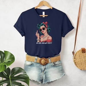 May include: A navy blue t-shirt with a graphic of a woman with red hair and sunglasses. The text "Is he dead yet?" is printed below the graphic.