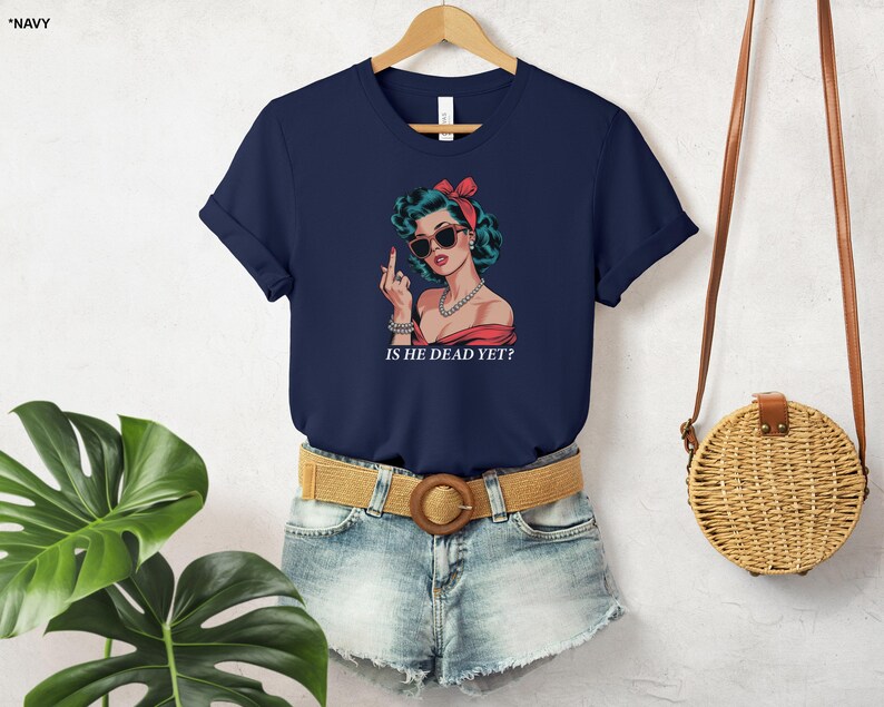 May include: A navy blue t-shirt with a graphic of a woman with red hair and sunglasses. The text "Is he dead yet?" is printed below the graphic.