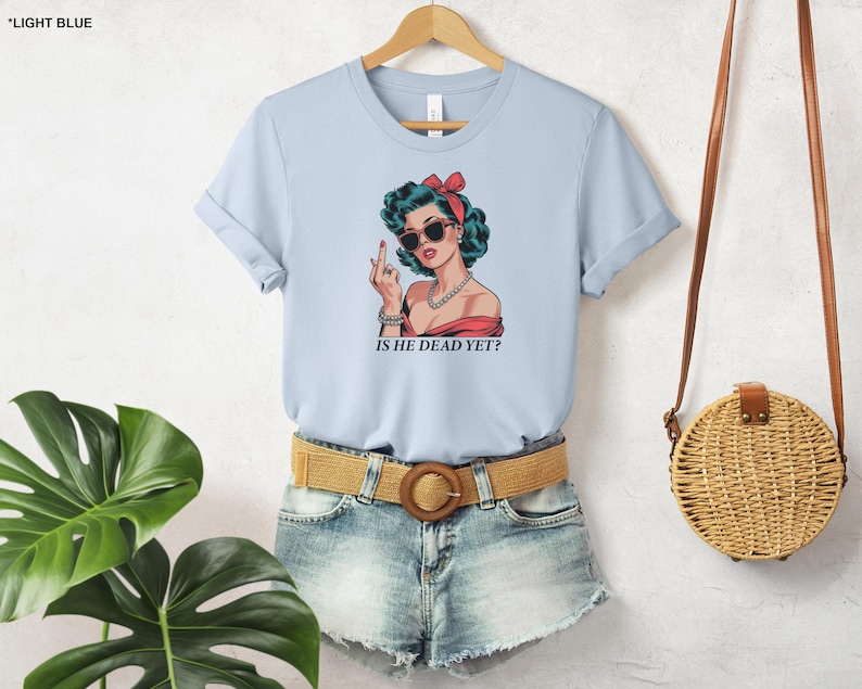 May include: Light blue t-shirt with a graphic of a woman wearing sunglasses and a red bow in her hair. The woman is giving the middle finger and the text "Is he dead yet?" is printed below the graphic.